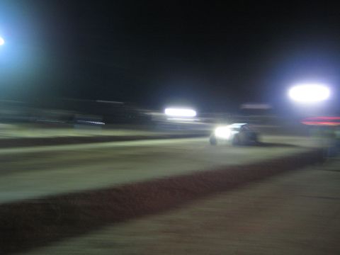 1990 Ford Bronco - race vehicle with bright headlights crosses a floodlit dirt track at night, captured in a heavily blurred panning shot.