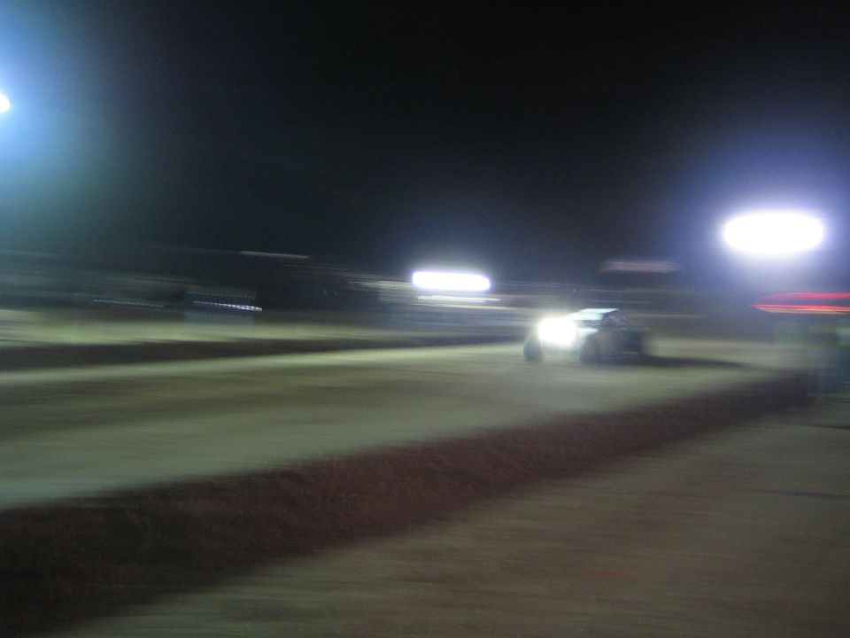Race vehicle with headlights crossing a floodlit dirt track at night, blurred