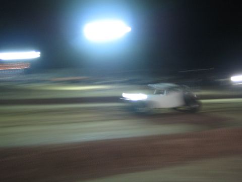 1990 Ford Bronco - blurred off-road race truck speeds across a dirt track at night, headlights on beneath bright stadium lighting.