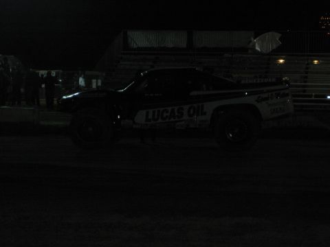 1990 Ford Bronco - 'Lucas Oil' sponsored off-road truck sits on a dirt course at night in front of dimly lit empty grandstands.