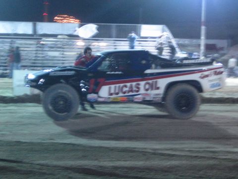 1990 Ford Bronco - red, white and blue #17 'Lucas Oil' off-road truck catches air over a dirt jump under night lighting as spectators watch.