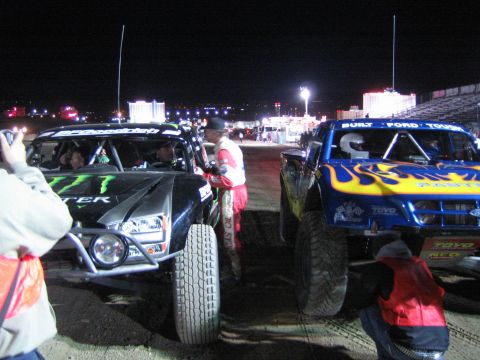 1990 Ford Bronco - Two trophy-style off-road trucks, one with Monster Energy graphics and one with a flame paint scheme, stage at night amid crew and a lit sk…