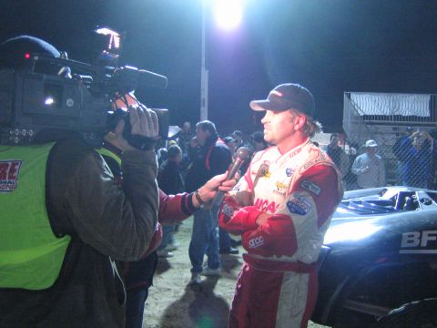 1990 Ford Bronco - driver in a red and white firesuit is interviewed beside a race truck at night while a TV cameraman films under bright lights.