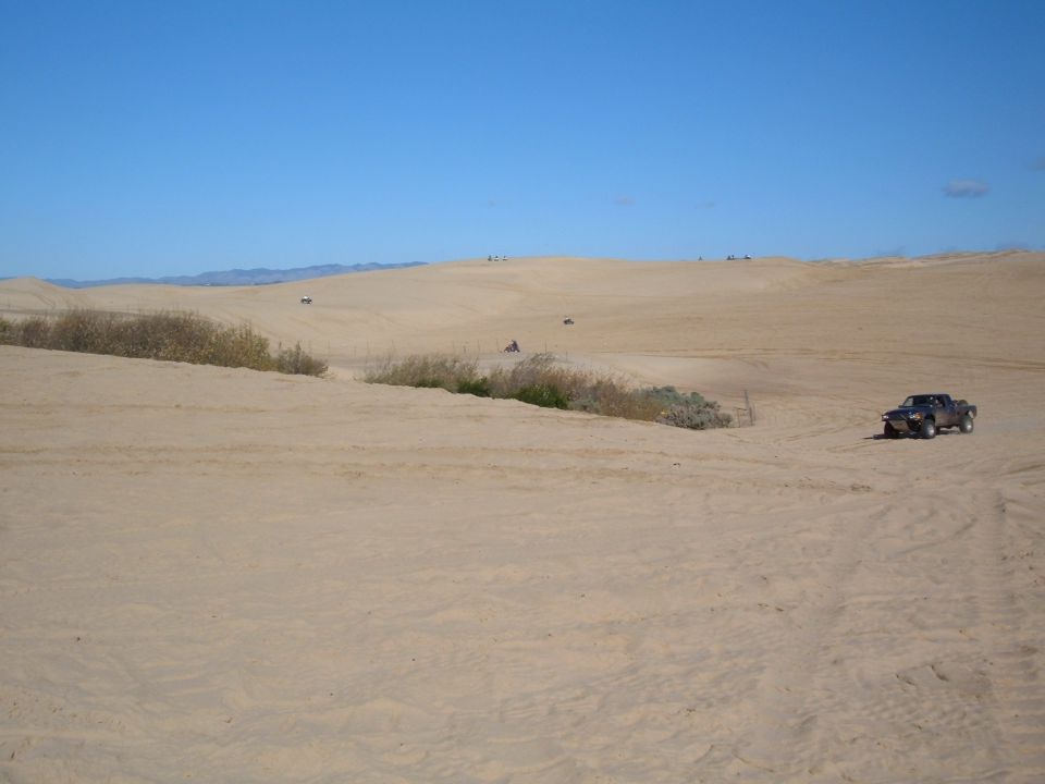 Dark colored pickup truck driving on a sandy dune under a clear blue sky. Other ATVs are visible in the distance