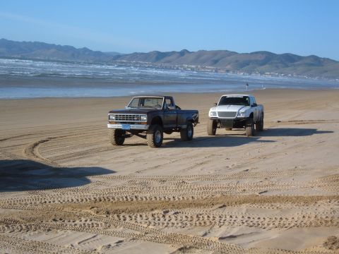 1985 Ford Ranger - Two pickup trucks drive on a sandy beach near the ocean with mountains in the distance.