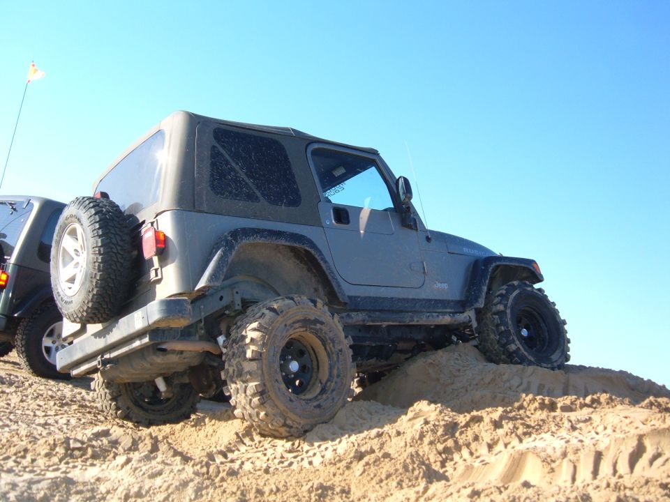 Gray Jeep Wrangler with large tires climbing a sand dune, another vehicle partially visible to the left