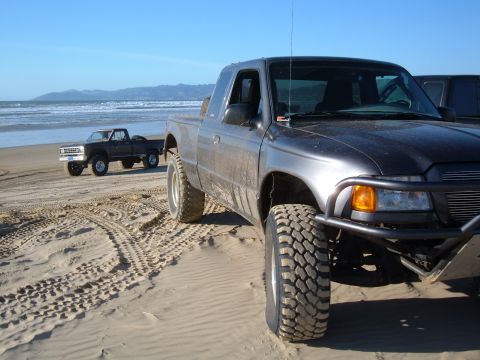 1985 Ford Ranger - Two pickup trucks are parked on a sandy beach with the ocean and hills in the background.