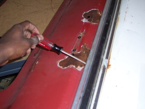 1978 Ford Bronco - A hand uses a screwdriver to probe rust holes on a red car door, with a Supermotors.net logo in the corner.