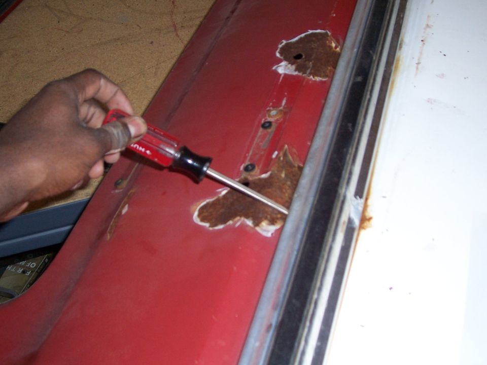 Red car door with rust holes being worked on by a screwdriver. A Supermotors.net logo is visible