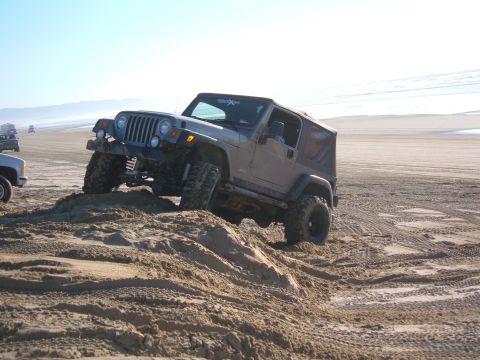 1985 Ford Ranger - A gray Jeep Wrangler TJ navigates a sandy incline on a beach, with other vehicles visible in the distance.