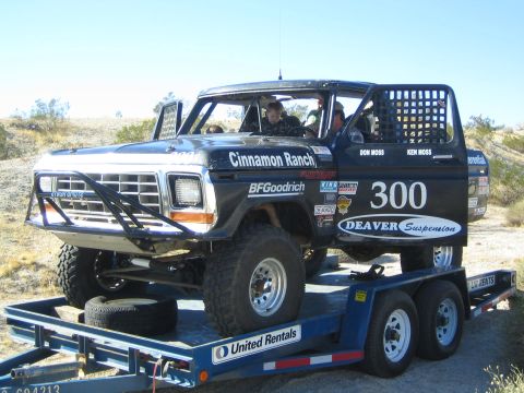 1990 Ford Bronco - black late-1970s Ford-style off-road race truck #300 lettered 'Cinnamon Ranch' sits chained to a United Rentals trailer in the desert.