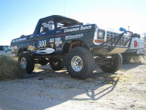 1990 Ford Bronco - black classic Ford off-road race truck #300 'Cinnamon Ranch' sits in desert brush on raised suspension, with another truck parked behind.