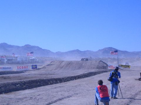 1990 Ford Bronco - Photographers watch from a dusty desert course as a distant off-road truck approaches a jump, with flags and sponsor banners along the trac…