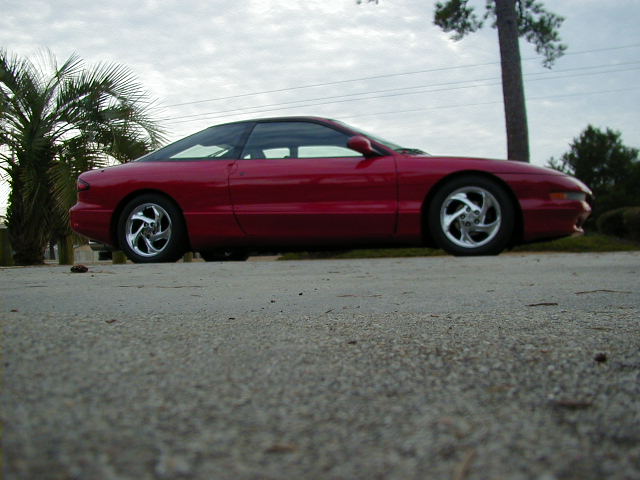 Side view of a red 1997 Ford Probe parked on pavement with palm trees and a utility pole in the background