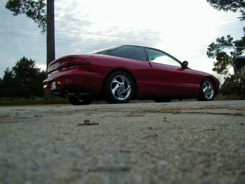 1997 Ford Probe - A red coupe with custom wheels is seen from a low angle, parked outdoors under an overcast sky.
