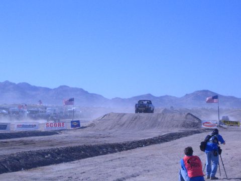 1990 Ford Bronco - off-road race truck crests a dirt jump on a dusty desert course lined with SCORE and BFGoodrich banners as photographers watch.
