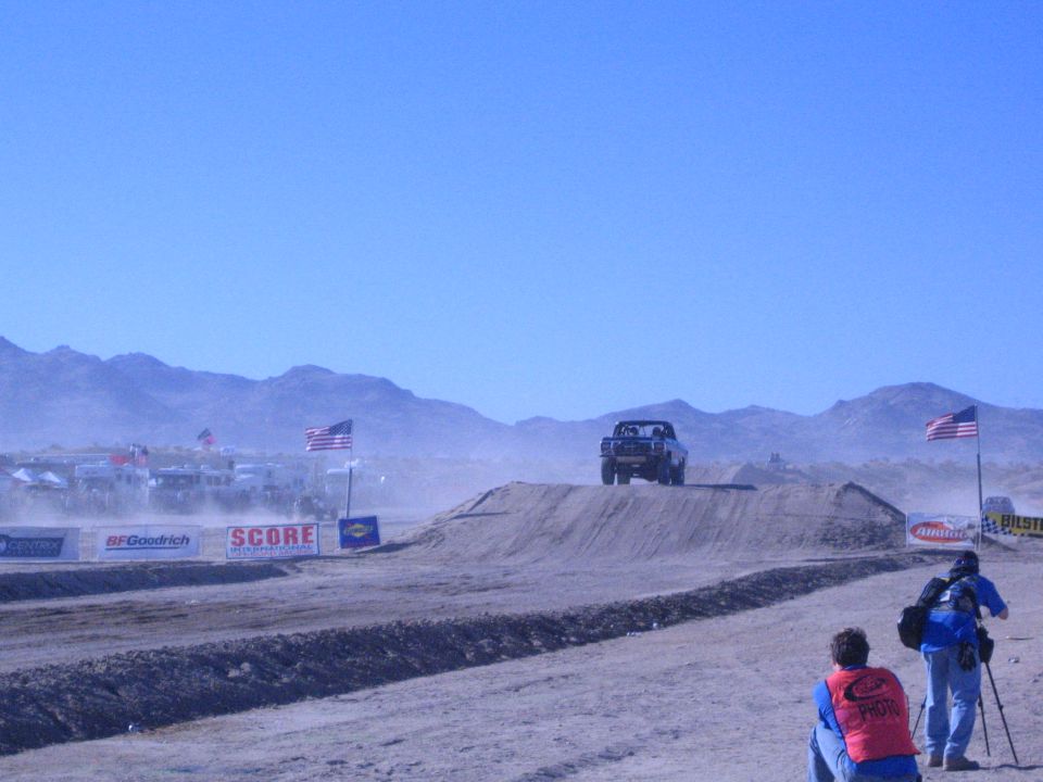 Off-road truck airborne over a dirt jump in a dusty desert course with flags and spectators