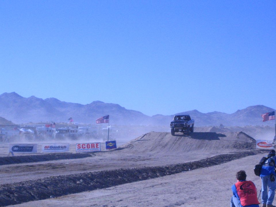 Dark off-road truck launching off a dirt jump on a dusty desert course with mountains behind