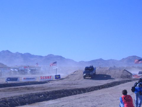 1990 Ford Bronco - off-road truck drives over a dirt berm on a dusty desert course backed by mountains, with flags flying and spectators watching.