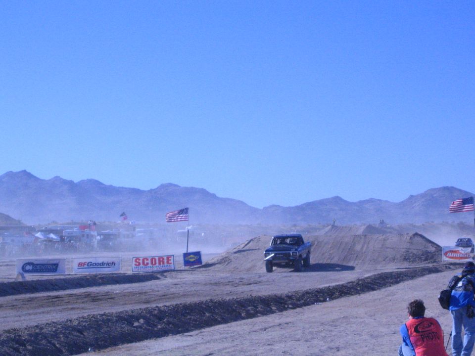 Blue off-road truck on a dusty desert dirt course with mountains, flags and a second truck