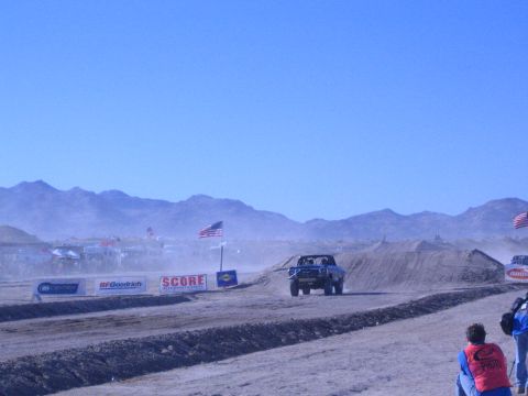 1990 Ford Bronco - blue off-road truck crests a dirt berm amid heavy dust on a desert course lined with American flags and a second truck visible behind.