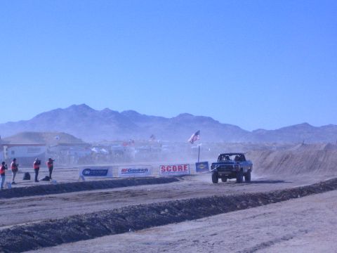1990 Ford Bronco - race truck kicks up dust on a dirt course while spectators in red vests stand behind sponsor banners under desert mountains.