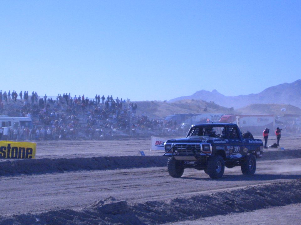 Blue lifted race truck number 300 on dirt course before packed grandstands in desert