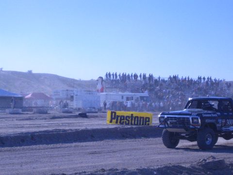 1990 Ford Bronco - blue race truck numbered 300 passes a yellow Prestone banner with a large crowd watching from the hillside grandstands.