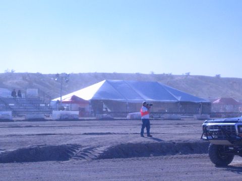 1990 Ford Bronco - person in a safety vest aims a camera on the dirt course near a large white tent, with the front of a lifted race truck at right.