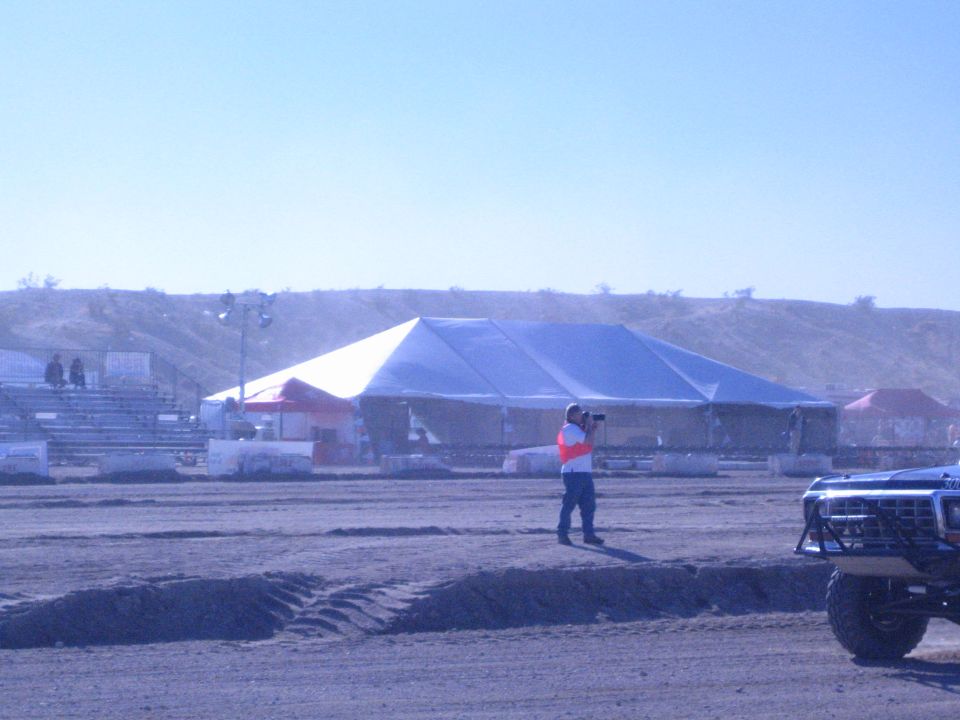 Person in safety vest with camera on dirt course beside large tent and a race truck front