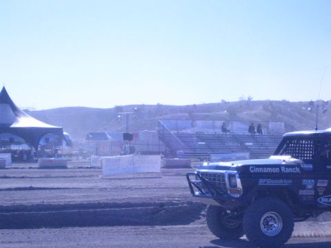 1990 Ford Bronco - blue 'Cinnamon Ranch' race truck sits front three-quarter on the dirt course beside a peaked tent and empty grandstands.