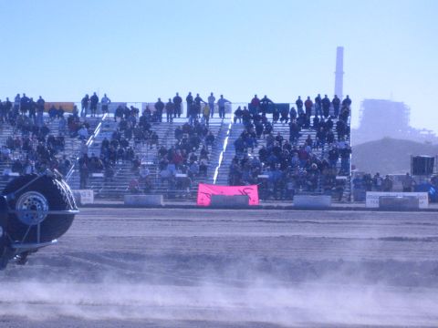 1990 Ford Bronco - Spectators fill metal grandstands behind a pink banner while dust drifts past a vehicle wheel in the foreground of the dirt arena.