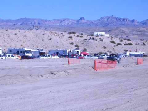 1990 Ford Bronco - desert race pit area lined with RVs, trucks and orange snow fencing spreads out below rugged mountains.