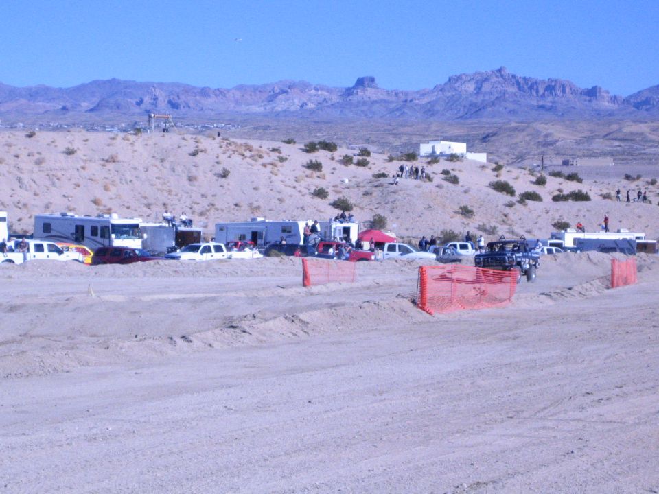Desert race pit area with RVs, trucks, orange fencing and rugged mountains behind
