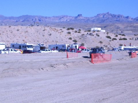 1990 Ford Bronco - wide dirt race course curves past RV pits and orange fencing in a broad desert valley ringed by mountains.