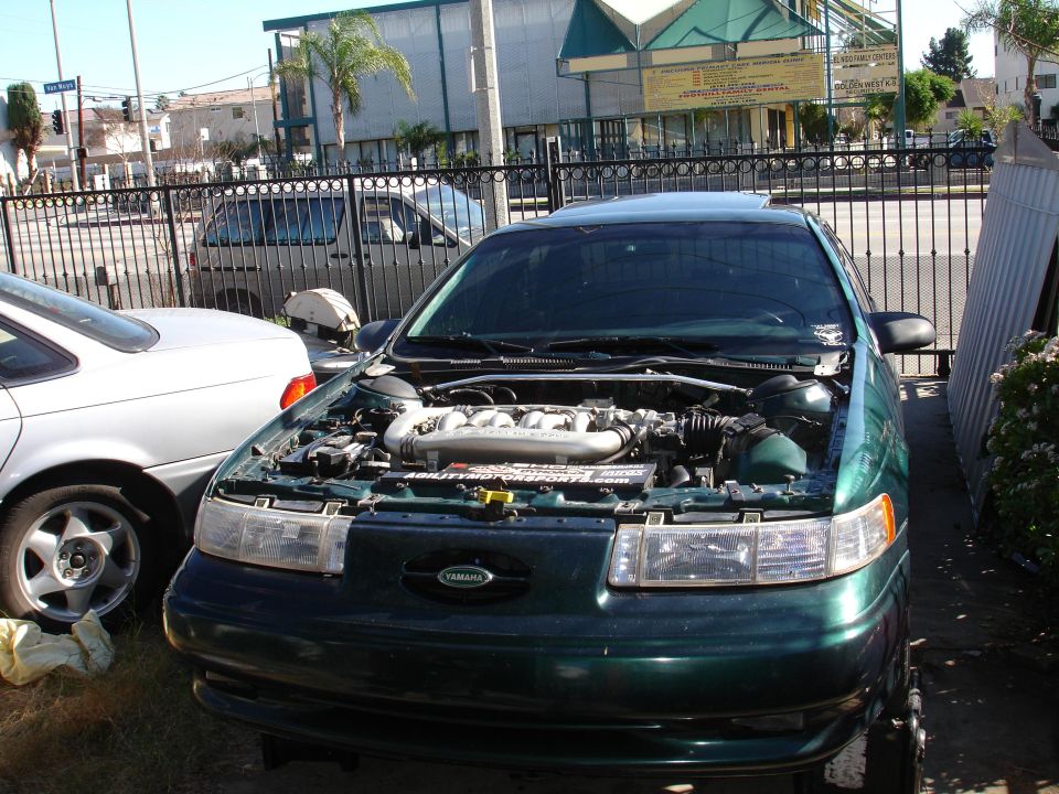 Green 1993 Ford Taurus, hood open, showing engine. Another car is parked to the left