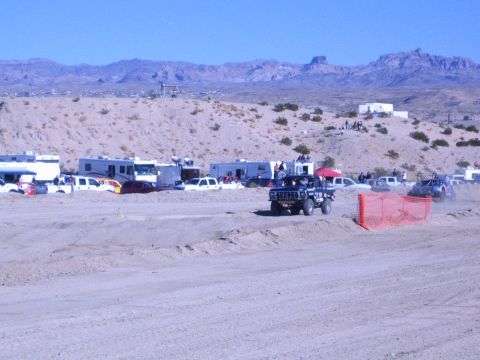 1990 Ford Bronco - boxy black off-road vehicle rounds a bend on the desert dirt course with RV pits and mountains behind.