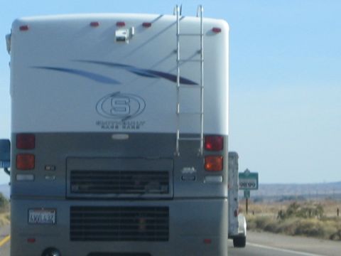 1990 Ford Bronco - rear of a silver Country Coach motorhome towing a trailer travels a desert highway, showing a California license plate.
