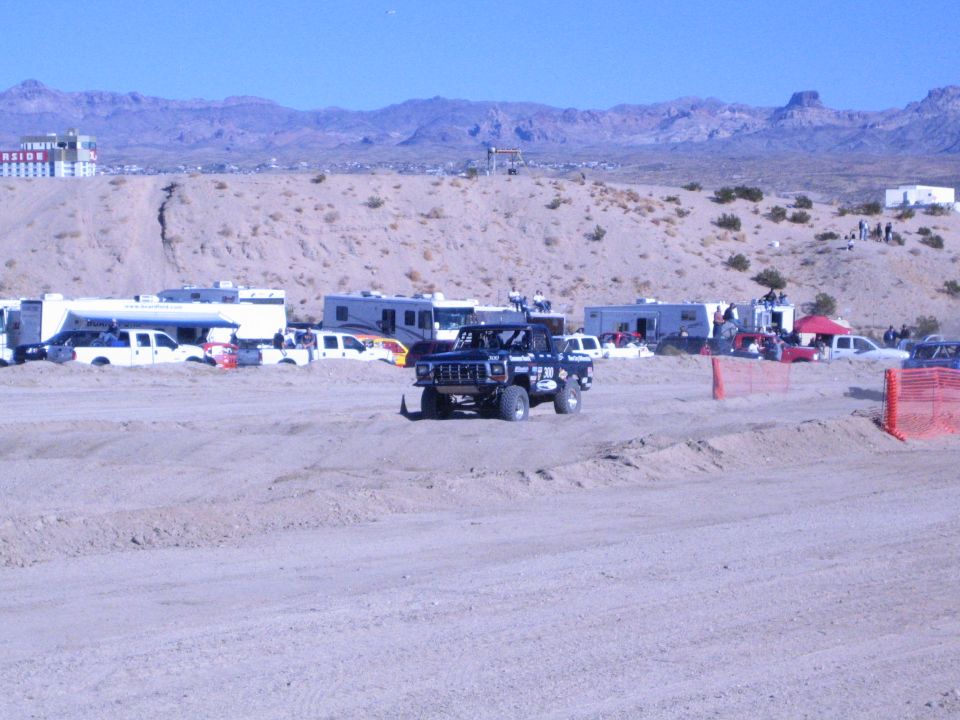 Black off-road truck on dirt course past RV pits and orange fencing in desert valley