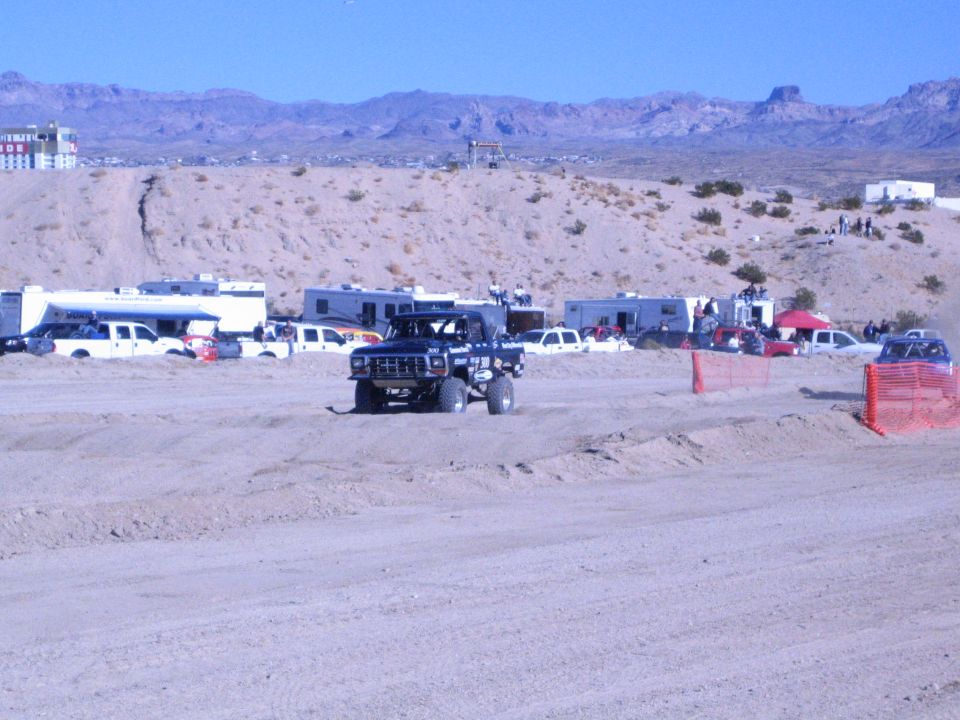 Dark off-road race truck on a dirt course with desert mountains and RVs behind