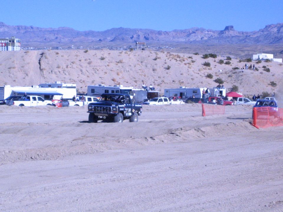 Lifted dark race truck on dirt track with parked RVs and arid hills behind