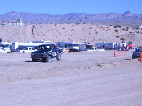 1990 Ford Bronco - dark off-road race truck crosses a dirt course with RVs and a red multi-story building in the desert behind.