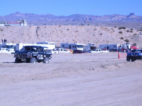 1990 Ford Bronco - dark Ford race truck and a second buggy on a dirt course with RVs and a red riverside building behind.