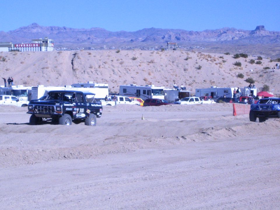 Two off-road trucks on a dirt course with RVs and red riverside building