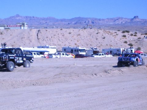 1990 Ford Bronco - Number 300 Ford race truck and a blue race truck on a dirt track lined with RVs under a clear desert sky.