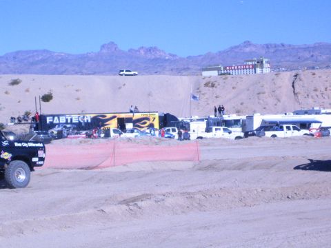 1990 Ford Bronco - Off-road race pits with a Fabtech banner, support trucks and trailers below sandy hills and a red riverside building.