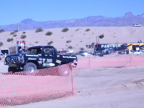 1990 Ford Bronco - Black Ford race truck number 300 with sponsor decals sits behind orange fencing in a desert pit area.