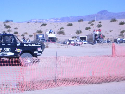 1990 Ford Bronco - rear of black race truck number 300 on a dirt course with spectator platforms and desert hills behind.