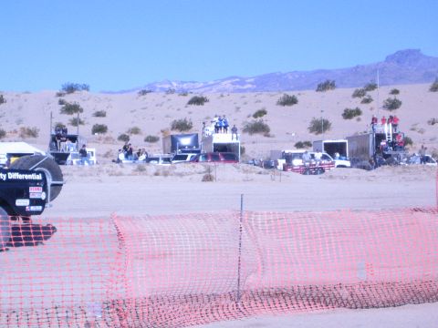 1990 Ford Bronco - desert race spectator and pit area with viewing platforms and support trucks behind orange fencing.