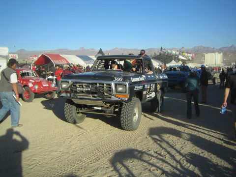 1990 Ford Bronco - Black Ford race truck number 300 rolls through a crowded desert pit area lined with tents and spectators.